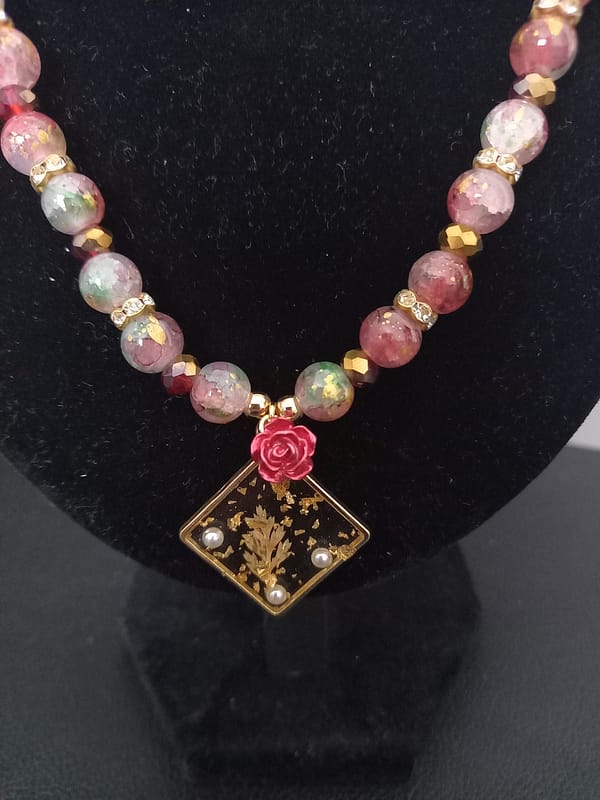 20250120_144742 Collier en perles marbrées craquelées rouges et vertes, séparées de strass et perles dorées, avec pendentif losange à feuille d’or et rose rouge.