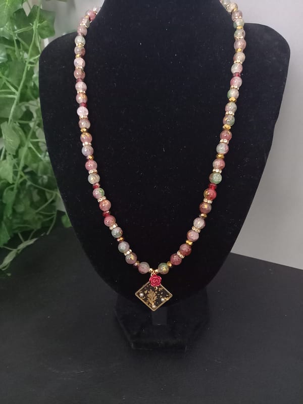 20250120_144804 Collier en perles marbrées craquelées rouges et vertes, séparées de strass et perles dorées, avec pendentif losange à feuille d’or et rose rouge.